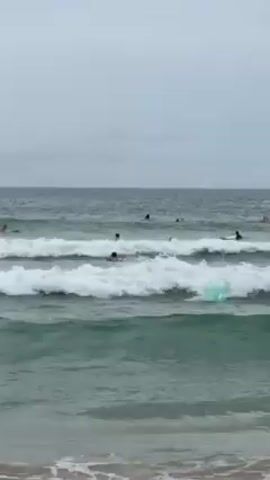 The video shows a beach with several people surfing in the ocean. The sky is overcast and the water is a greenish color with white waves. People are spread out in the water, some riding waves and others paddling. A green surfboard is visible near the front of the scene.