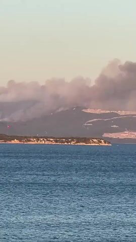 The video shows a wide shot of a body of water in the foreground, with a coastline visible in the distance. On the coastline, there is a peninsula with trees and a red flag. Behind the peninsula, a mountain range is visible, with plumes of smoke rising from it, indicating a wildfire. The sky is clear and appears to be sunset or sunrise, with soft lighting. The overall scene depicts a natural landscape impacted by a fire.