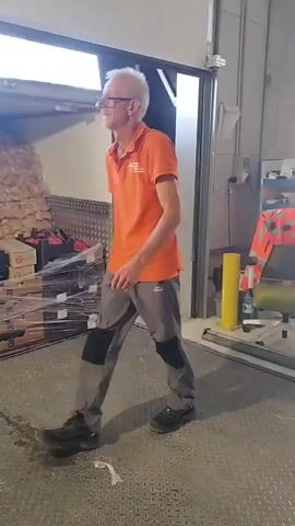 A man wearing an orange shirt and grey pants walks in front of a loading dock. The man has glasses and white hair. He is walking towards the right of the screen. Behind him is a truck, filled with boxes and what appears to be insulation.