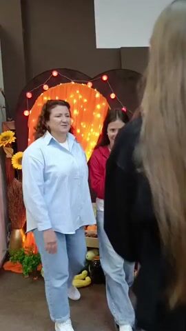 A woman with curly brown hair wearing a light blue striped button-up shirt over a white t-shirt and light blue pants is standing and looking to her left. She is in an indoor setting with a dark brown wall behind her. To her left, there is a decorative arch with orange fabric draped inside, illuminated by many small orange lights. Red lights are strung along the top of the arch. Two sunflowers are visible to the left of the arch, and a wooden cart filled with various fruits and vegetables is positioned in front of the arch. On the ground in front of the cart, there are several jars and more fruits and vegetables. The floor appears to be carpeted. Another person's back, with very long brown hair, is visible in the foreground to the right.