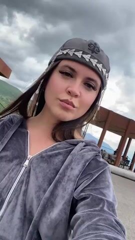 A medium shot video shows a young woman in a gray velour tracksuit and a gray hat adorned with a silver pattern and chain details on the sides. She is holding the camera and appears to be taking a selfie. She has dark hair and is wearing makeup, including dramatic eyeliner and false eyelashes. She is outdoors, and the background suggests a mountainous or hilly landscape. Behind her, there is a modern building with large windows and some outdoor seating areas. The sky is cloudy. The camera pans slightly, offering glimpses of the surrounding scenery.