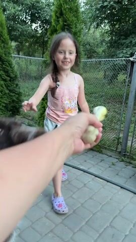A young girl with a pink top and denim skirt is laughing while playing with a dog in an outdoor, paved area with greenery in the background. A person's hand is visible, holding a yellow object, possibly a toy or treat, which the dog is interacting with. The girl is in motion, with her arms raised, and the dog is also blurred due to movement.