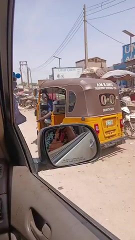 The video captures a bustling street scene in Badiko, Nigeria, under a bright, sunny sky. The footage is filmed from inside a vehicle, with its side mirror reflecting a partial view of the interior.

The primary subject is a yellow and brown Keke Napep (auto-rickshaw) prominently featuring "ANKAWA TRANSPORT SERVICES" and a phone number "08108742950" on its rear. A man in a blue patterned shirt is seen driving the Keke, initially looking towards the camera before turning his gaze to the side. A sticker with the number "12" is visible on the Keke's side.

The street is unpaved and dusty, indicating an active market or commercial area. Various individuals are visible: some are walking, including a woman in a black hijab and colorful dress, and a man in a red top; others are seated under large market umbrellas. Market stalls with goods, blue barrels, and a parked motorbike contribute to the lively atmosphere. Commercial buildings line the street, with prominent signs displaying "UNIFOAM ALMAD GROUP" and another sign possibly reading "EUROFOAM." A white SUV and two wooden handcarts, one marked with "NO PARKING" on a bucket, are also part of the scene.