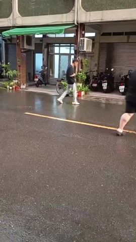 The video shows a wet street scene in what appears to be a residential or commercial area. The street is marked with a yellow line. Two people are seen walking along the street. Multiple parked scooters and cars are visible along the sides of the street. The buildings lining the street are mostly two or three stories high and appear to be a mix of residential and commercial properties. The weather is rainy, and the street surface is wet and reflective.