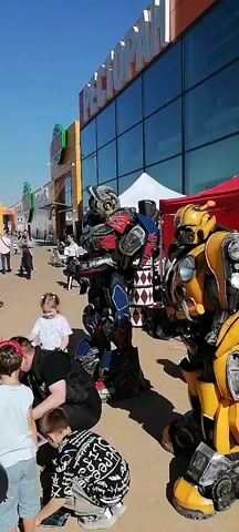 Two individuals in elaborate robot costumes resembling Optimus Prime (red, blue, and black) and Bumblebee (yellow and black) from Transformers are standing outdoors on a paved area. The Optimus Prime robot performs a slight arm movement. In the lower left foreground, a woman in a black shirt is bent over, interacting with two young children. One child, a girl in a white and blue striped shirt, is standing near the woman. In the background, other people are walking along a street lined with buildings that have large, reflective glass windows and commercial signs, under bright sunlight. A red tent and speakers are visible behind the Bumblebee robot on the right.