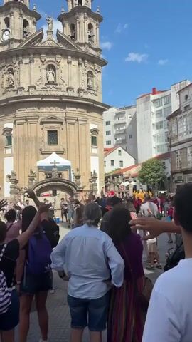 The video shows a street festival in front of a large stone building with two towers. A crowd of people is gathered, many of whom are facing the building and raising their hands. Some people are wearing casual clothing, while others are dressed in festive attire. There are tents set up in front of the building, and a white cross is visible on one of them. The sky is blue with scattered clouds, and the sun is shining. The overall atmosphere appears to be celebratory and lively.