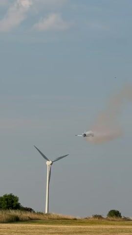 A remote-controlled jet airplane is seen flying across a clear sky, emitting a trail of white smoke. The plane is white with red and blue accents. In the foreground, a large wind turbine stands tall, with its blades appearing to be stationary. The surrounding landscape consists of dry, yellow grass and a few green bushes. Power lines and pylons are visible in the background, suggesting an open, rural environment. The video captures the plane in mid-flight, performing an aerial maneuver.