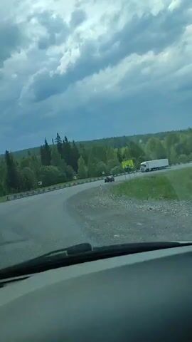 The video shows a view from inside a car driving on a paved road in a somewhat rural area. The sky is cloudy with patches of blue. Green trees line the side of the road. In the distance, there is a slight elevation visible. A large semi-truck and a white van are visible on the road ahead. A sign is visible on the right-hand side of the road, next to a turn. The road curves to the left. Music is playing in the background.