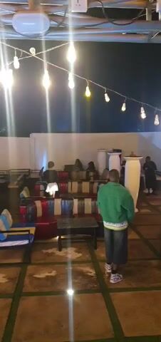 The video shows an outdoor patio area at night, illuminated by string lights. Several people are scattered around, some seated on colorful sofas and chairs, others standing. A woman in a black jacket and white skirt is prominently featured walking across the patio. A man in a green jacket walks away from the camera. The area appears to be a lounge or seating area with tables and various decorative elements.