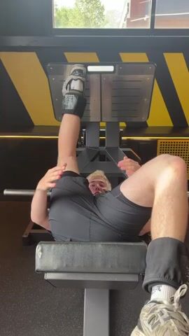 A person is performing a leg press exercise on a gym machine. The person is lying on their back on a padded bench with their feet positioned on the footplate of the leg press machine. They are wearing black shorts and ankle weights on both legs. The leg press machine is gray and yellow. The person is pushing the footplate away from them with their legs, extending them fully. The environment appears to be a gym with yellow and black patterned walls in the background.
