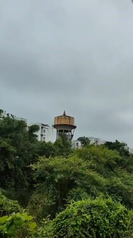 The video shows a water tower with a red flag on top, surrounded by lush green trees and vegetation. In the background, there is a white building. The sky is overcast, suggesting a cloudy or rainy day. The camera pans across the scene, highlighting the water tower, trees, and building.