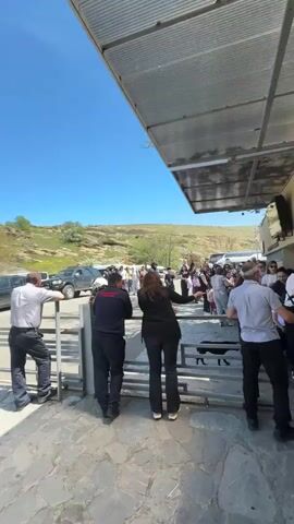 The video shows a group of people standing outside near a gate and souvenir shop, with the Georgian flag waving in the background on a clear, sunny day. Some people are waiting near a metal gate, while others are gathered under the shelter of the souvenir shop, looking at merchandise and each other. Beyond the group, a grassy landscape with rocky formations and trees can be seen. The camera pans across the scene, capturing different viewpoints. The ground is paved with stone. Several cars are parked near the gate.