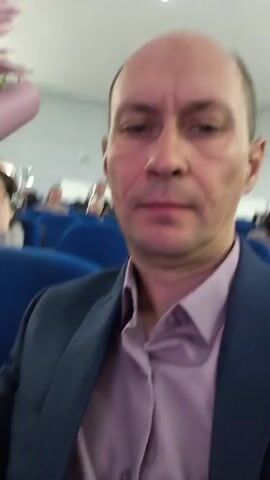 A man with a shaved head, wearing a dark blue suit jacket over a light purple collared shirt, is filming a selfie video in an auditorium. He is seated in a blue upholstered chair. To his left, a bouquet of pink and white lilies wrapped in light purple paper is held by a hand, presumably his own, though the hand is mostly out of frame. Behind the man, other audience members are visible, seated in rows of similar blue chairs. In the background, there is a stage area with lighting and some architectural features. The man blinks in the first few seconds of the video.