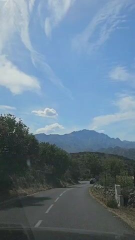 A car is driving down a winding asphalt road with white lines. The road is bordered by trees and rocks. In the distance, there are mountains under a blue sky with white clouds. The view is from inside the car, as indicated by the windshield and dashboard in the foreground. The time code indicates that this is the beginning of the video.