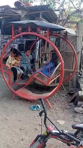 A group of four children are playing on a red circular swing set in an outdoor area. Two boys are seated on one side of the swing, one boy is seated on the other side, and a girl is seated on the opposite side of the girl. The children appear to be happy and engaged in playing together. The swing set is made of metal and has a roof over the top. The ground around the swing set is dirt. There is a bicycle in the foreground of the image. The background of the image shows some buildings and trees.