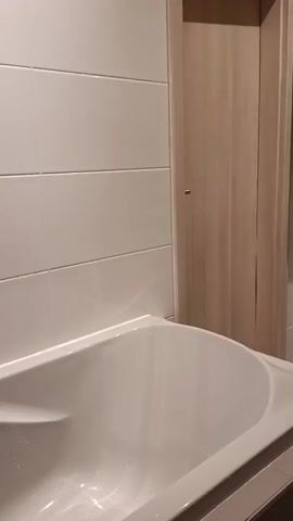 This is a video tour of a bathroom in Roßlau, Germany. The bathroom appears to be located in an attic space, as evidenced by the sloping ceiling and exposed wooden beams. 

The room contains a white bathtub, a sink with a cabinet underneath, a toilet, a washing machine, and a wall-mounted cabinet with its doors open, revealing shelves with items on them. There is also a mirror above the sink, a towel rack with clothes draped over it, and a paper bag on the floor. The walls are tiled in white and painted yellow. The flooring appears to be grey tile.

The video begins with a shot of the bathtub and then pans to show the rest of the bathroom, including the sink, toilet, washing machine, and cabinets. The camera moves smoothly, providing a clear view of the space. There are no people visible in the video. The lighting suggests it is daytime, and there are no clear indicators of the weather outside. The overall impression of the bathroom is functional and lived-in, with laundry visible on the rack and clothes on top of the washing machine. The open cabinet suggests recent use or preparation for use.