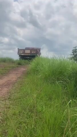 The video shows a view of a large, rusty dump truck parked on a dirt path. The truck is facing away from the camera and appears to be on a grassy hill with lush green grass on either side of the path. The sky is cloudy with dramatic cloud formations. The path leads away from the truck towards the foreground of the video, where it is partially obscured by the grass. There are no people visible in the video. The overall scene suggests an outdoor setting, possibly rural or undeveloped, with the truck appearing to be abandoned or parked for an extended period.