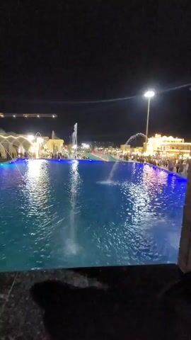 This media file captures a nighttime scene at a location in Satkak, Uzbekistan, likely an entertainment venue or park. The primary subject is a large, illuminated swimming pool or water feature. The water is a vibrant blue, reflecting the surrounding lights and the dark night sky. Several fountains are active in the pool, creating dynamic water patterns.

In the background, a crowd of people are visible, suggesting a public gathering or event. They are gathered along the edge of the water feature and near buildings with illuminated facades. The architecture of the buildings appears somewhat ornate, with distinct lighting creating a festive atmosphere. To the left, a construction crane is visible against the night sky, hinting at ongoing development or a nearby project. The lighting is predominantly artificial, with bright spotlights and colored lights illuminating the area.

The time of day is clearly night, with a deep dark sky. There are no indicators of weather conditions, but the overall scene suggests a pleasant evening, as indicated by the numerous people out and about. The mood conveyed is one of lively entertainment and enjoyment, with the bright lights and active water features contributing to a festive ambiance. No discernible text is visible within the frame.