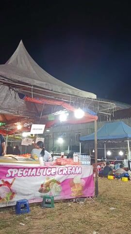 This media file captures a lively night scene at an outdoor market or event, likely in Sambirata, Indonesia, given the context. The primary focus is an ice cream stall, brightly lit by overhead lights and adorned with a vibrant pink banner that reads "SPECIAL ICE CREAM" in large, bold white lettering. The banner also features images of ice cream sundaes and text like "Special Delicious" and "Best".

Several people are visible within the frame. In the foreground, a person is seen at the ice cream counter, appearing to be serving or interacting with customers. Behind the counter, more people, possibly customers or stall workers, are gathered. Further in the background, beyond the ice cream stall, a group of people are seated, suggesting they are enjoying the event or perhaps waiting for something. They are positioned in front of tents and buildings, under artificial lighting, indicating it is nighttime.

The setting appears to be an open, grassy area with some dry grass and dirt. The atmosphere seems to be one of a community gathering or fair, with the illumination and the presence of multiple people suggesting a festive or social occasion. The weather appears to be clear, given the outdoor setting and the presence of people. The overall impression is a bustling evening marketplace with a popular ice cream vendor at its center. The visible text confirms the nature of the business and hints at the quality of the product.