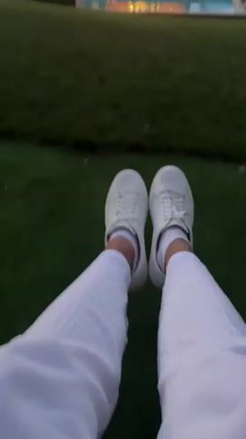 The video opens with a close-up of a person's white sneakers and white pants, their legs crossed. The background is blurred, showing a grassy lawn, a swimming pool, and a house with lights on and string lights draped across the patio. The camera pans slightly, showing the person's legs shifting and their feet moving. A faint audio track plays, featuring music and indistinct voices. The video continues to show the person's legs and feet against the backdrop of the house and pool area. The scene appears to be outdoors at dusk or evening.