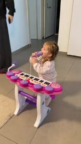 A small child with pigtails stands at a toy keyboard/drum set. The keyboard is pink, white, and purple, with a small black-and-white keyboard. The child wears a striped white-and-pink pajama top and white pants. She holds a purple microphone up to her mouth and appears to be singing or making sounds into it. A person in dark clothing holds a red object in the background. The floor is tiled. A beige rug is visible. A doorway is visible behind the child.