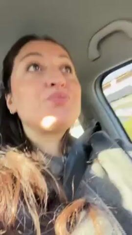 A woman with long brown hair and wearing a gray sweatshirt is sitting in the driver's seat of a car. She is wearing a seatbelt. She looks at the camera and then looks at the road as she drives. The weather outside is sunny. Music is playing.