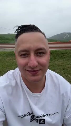 A man with short dark hair, styled with a side part and gelled on top, is filmed from the chest up. He has light skin, blue-green eyes, and a slight smile. He is wearing a white t-shirt with a black graphic design on the lower left side. The background appears to be an outdoor setting with green grass in the foreground, a blurred orange barrier in the mid-ground, and green, tree-covered hills with some snow on top in the distance. The sky is overcast and gray. The man is looking directly at the camera.