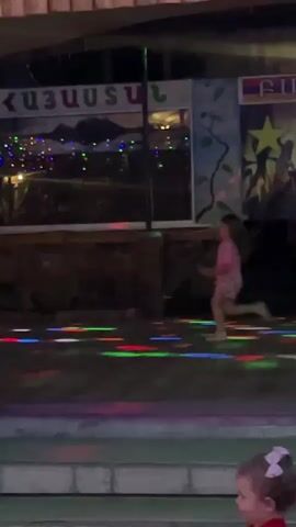A young girl in a pink shirt and light-colored shorts is dancing on a colorful dance floor. The dance floor is illuminated with various colored lights. In the background, there is a large sign with writing in a foreign language and a mural depicting a landscape. There are also strings of lights hanging above. A man wearing an apron walks into the frame from the left and descends a set of stairs. Further down the stairs, another young child in a red shirt and patterned pants is standing. The environment appears to be an outdoor or semi-outdoor venue, possibly a restaurant or entertainment area, with wooden structures and ambient lighting.
