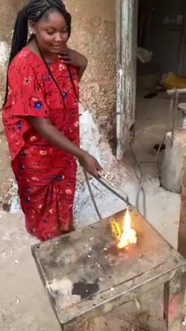 A woman stands outdoors by a small, dirty metal table holding a blowtorch that is pointed at a metal object on the table, with flames coming from the object. The woman has dark skin and long, dark braids, and she is wearing a long red dress with a floral pattern. She looks down at the object and has one hand raised to her chest. The scene appears to be outside a building with a doorway slightly visible behind her. There is a metal tank standing to her right.