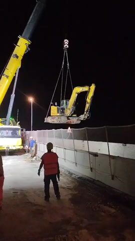 A yellow excavator is suspended in the air by a yellow crane at night. The excavator is hanging from a hook with cables. The crane is partially visible on the left side of the frame. In the foreground, two construction workers are standing. One worker is wearing an orange uniform and the other is wearing a red vest and a yellow helmet. Another worker, wearing a blue shirt, is visible in the background near a truck. There is a concrete barrier with a fence on top of it behind the workers. The ground is covered in dirt and gravel. Streetlights and a dark sky are visible in the background.