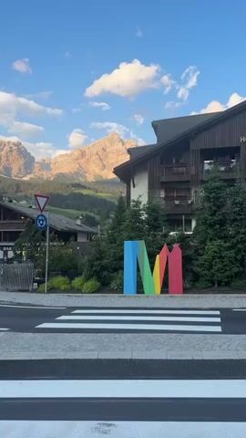 The video shows a roadside scene with a pedestrian crossing in the foreground. Behind the crossing, a large, colorful sculpture of the letter "M" stands. Further back, a street curves to the left, bordered by a wooden fence. Beyond the fence, there are buildings, including one with a sign that says "PIZZERIA BAR". Trees are visible throughout the scene. In the background, a lush green mountain slope leads up to a dramatic mountain range with rocky peaks, bathed in the warm light of the setting sun. The sky is blue with scattered white clouds. The overall impression is a picturesque mountain village setting.
