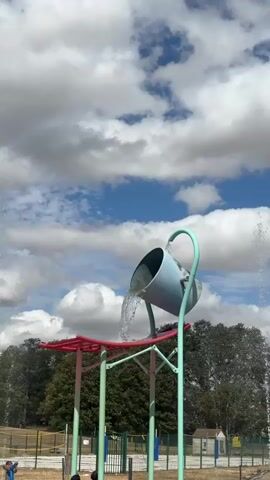 The video shows an outdoor water play area with a large blue bucket tipping over and releasing a large amount of water. The bucket is suspended from a green pole with a curved arm. Beneath the bucket is a red and pink structure with green poles. The sky is blue with fluffy white clouds. In the background, there are trees, a fence, and some small buildings. Two children are visible in the foreground, one on the left and one on the right, appearing to be playing in the water. The child on the left is wearing a dark swimsuit and is bending over. The child on the right is wearing a light-colored swimsuit and is standing. Water is spraying from various points around the play area. The overall scene depicts a sunny day at a water park or splash pad.