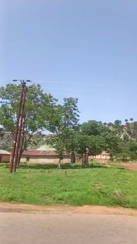 The video shows a drive-by view of a rural landscape on a clear, sunny day. The camera is likely inside a moving vehicle, and the focus is on the scenery passing by. The scene includes a mix of vegetation, such as trees and grassy areas, along with simple, possibly brick, buildings or houses. In the distance, there is a rocky, hilly area dotted with trees. Utility poles and wires are visible alongside the road. The landscape features red soil or dirt. The overall impression is of a quiet, rural setting.