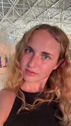 A young woman with blonde, wavy hair and blue eyes is filmed in a close-up selfie. She is wearing a black tank top. The background appears to be an indoor climbing gym with a high ceiling, exposed metal beams, and climbing walls visible in the distance. The lighting is bright and even, suggesting artificial illumination. The woman is looking directly at the camera with a slight smile.