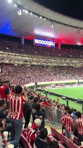 The video shows a soccer game at a packed stadium. The stands are full of fans wearing red and white striped jerseys, many of whom are holding up their phones to record the event. A large screen in the background displays the word "GOOOOOL!". The field is green and well-lit, and players in light blue uniforms are visible. Security personnel stand near the edge of the field. The stadium's lights alternate between red and blue.