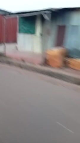 The video shows a quick street-level perspective. It appears to be filmed while moving, possibly from a vehicle, passing by a series of buildings and structures along a street. The buildings are of various sizes, colors, and materials, including brick, cement, and corrugated metal. One structure displays a mural. A person wearing blue pants walks by. The street is paved, and there's a shallow drainage ditch alongside it. The sky is overcast. The video quality is blurry due to the speed and motion.