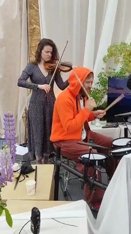 This video captures a live musical performance with multiple musicians. In the foreground, a person wearing an orange hooded sweatshirt and brown pants plays a drum set with drumsticks. To the left, a woman with brown hair, wearing a dark floral-patterned dress, plays the violin. Behind them, a person with dark hair and a black jacket plays a red bass guitar. Further in the background, two people sit on the floor playing acoustic guitars and singing into microphones. One of them is wearing a white garment, and the other is wearing a red headscarf. The environment appears to be indoors, with white drapes or curtains in the background, along with some greenery and a large picture of a person. There are also various audio equipment such as speakers and microphones visible.