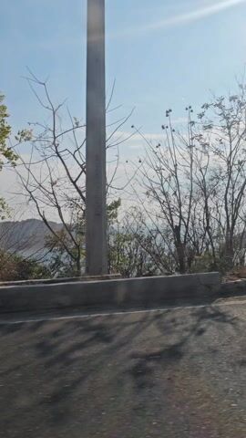 The video is shot from a moving vehicle, driving on an asphalt road. A short concrete barrier separates the road from a view of a bay with mountainous islands in the distance. The sky is blue with scattered clouds. A white car passes in the opposite direction. Bare trees and brush are visible along the roadside. Utility poles and power lines are also seen along the road. The video appears to be taken during the daytime in a coastal environment.