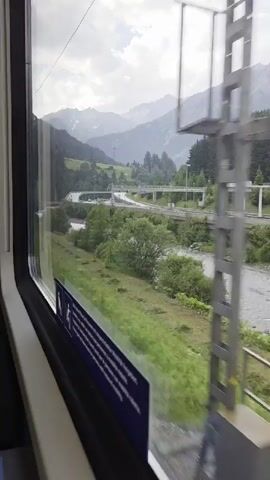The video captures a scenic view from inside a train traveling through a mountainous landscape, likely in Flirsch, Austria. The scene is dominated by lush green vegetation, a winding river, and majestic, cloud-kissed mountains in the background. The time of day appears to be overcast, suggesting either early morning, late afternoon, or a generally cloudy day.

From the train window, we see a river flowing alongside the tracks, bordered by dense, verdant greenery and trees. Beyond the river, a road with bridges and traffic, possibly a highway, curves through the valley, indicating human infrastructure within the natural setting. Power lines and poles are visible, further reinforcing the presence of civilization. The mountains themselves are a striking feature, with some peaks possibly capped with snow, shrouded in mist, contributing to a serene and slightly dramatic atmosphere.

Inside the train, a blue and white informational sticker is affixed to the lower part of the window frame. This sticker appears to contain text and icons, possibly indicating safety information, instructions, or accessibility features within the train carriage. The overall mood conveyed is one of peaceful travel and appreciation for the natural beauty of the mountainous region. The weather is subdued, with a soft, diffused light, enhancing the moody and tranquil ambiance of the landscape.
