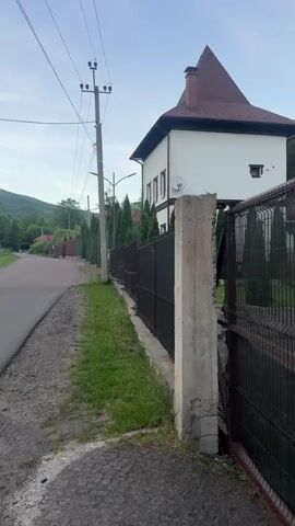 The video shows a view from a sidewalk along a residential street. On the right is a dark metal fence with concrete posts, beyond which stands a white house with a brown roof. Power lines and poles are visible on the left side of the street. In the background, green mountains can be seen under a slightly overcast sky.