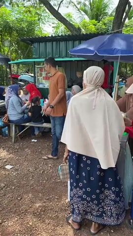 A man in an orange polo shirt and blue jeans walks past a food stall. Several people are seated at tables in front of the stall, including women and children. A blue umbrella is set up over one of the tables. The ground is covered in dirt and small stones. Trees and foliage are visible in the background, along with parked motorcycles.