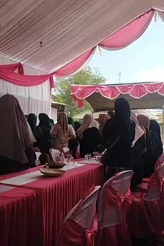 A gathering of women wearing hijabs are seen under a white tent decorated with pink fabric. Tables are set with pink tablecloths and chairs are draped with pink sashes. Some of the women are standing and talking, while others are seated. There are dishes of food and drinks on the tables. The overall atmosphere appears festive and celebratory.
