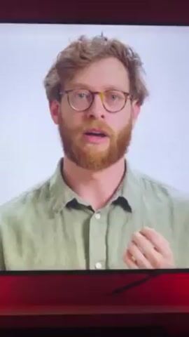 A medium close-up shot depicts a man with wavy brown hair and a beard, wearing round tortoiseshell glasses and a light green button-down shirt. He is gesturing with his hands as he speaks, his expression earnest. The background is a plain, bright white. The video is being displayed on a television screen, as indicated by the red border around the image.