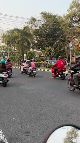 The video shows a street scene with several motorcycles and a car. In the foreground, a motorcycle with two riders is visible, and the motorcycle is emitting a significant amount of white smoke from its exhaust. Other motorcycles are on the road, some with riders and some appearing to be empty. In the background, a white car is traveling on the road, and beyond that, there are trees and buildings. A tall industrial chimney with dark smoke billowing from it is visible in the distance. The road appears to be paved asphalt, with some wear and tear visible. The overall lighting suggests it is daytime, possibly late afternoon. The camera is positioned at a low angle, looking forward along the road.