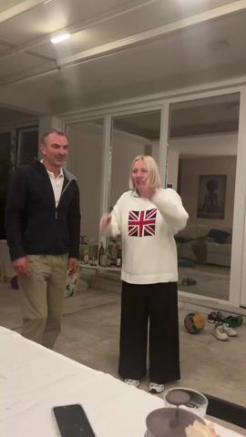 A man and a woman are in a brightly lit room with large glass sliding doors leading to another room. The man is on the left, wearing a navy blue jacket over a white shirt, and khaki pants. He’s looking at the woman and smiling. The woman is wearing a white sweater with a Union Jack flag on the front, and black wide-leg pants. Initially, she has her hands on her head, then she pulls the sweater up over her head and holds her arms out to the sides. There’s a table in the foreground with a cell phone on it. In the background, behind the glass doors, is a living room with a couch and a framed picture on the wall. There's also a table with liquor bottles and some other items on it. A soccer ball, shoes, and sandals are on the floor in the background.