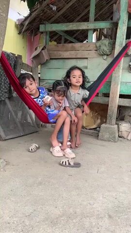 Three young girls are sitting in a red and black hammock. The girl on the left is wearing a blue and white t-shirt and is looking at the camera. The middle girl is wearing a gray and white t-shirt and blue shorts and is holding a white object. The girl on the right is wearing a green dress and is looking to the side. They are in an outdoor setting with a concrete ground. Behind them is a wooden structure with a thatched roof. There are also some sandals on the ground.