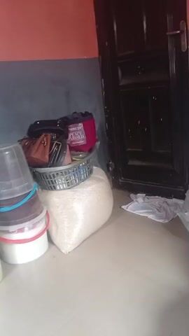 The video shows a collection of stacked buckets, a large bag of rice, and several purses in an indoor setting with red and gray walls. The objects are piled near a closed dark brown door and a gas cylinder.