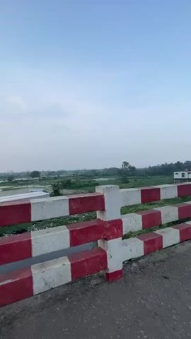 The video begins by showing a red and white striped guardrail along the side of a road, with a lush green landscape in the background. A motorcycle is being ridden, as evidenced by the rearview mirror reflecting the rider and the front of the motorcycle. The road appears to be an elevated bridge, as a body of water or low-lying land is visible below the guardrail. The sky is clear and blue with a few wispy clouds. As the video progresses, other vehicles become visible on the road, including a rickshaw and a bus. The motorcycle continues to travel forward, passing by the other vehicles. The scene is set in what appears to be a tropical or subtropical region, given the vegetation. The overall impression is one of transit through a semi-urban or rural area.
