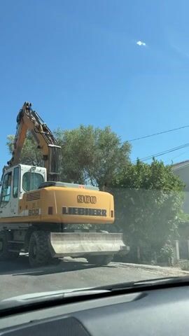 A yellow and white wheeled excavator is parked on a street. The excavator has the brand name "LIEBHERR" printed on its arm and body. A person is visible inside the cab of the excavator. The scene is outdoors with a clear blue sky and green trees in the background. The video appears to be filmed from inside a vehicle, with the dashboard and windshield visible in the foreground.