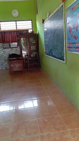 The video shows a classroom with a teacher interacting with students. The room has green walls and a tiled floor. A blackboard is mounted on one wall, and a world map is visible on another. The teacher, wearing a patterned shirt and black pants, is shown bending over to speak with students sitting at wooden desks. The students, all wearing green shirts, are engaged in activities at their desks. Natural light enters the room through windows with blinds. A projector is mounted on the ceiling. A wooden cabinet and a clock are also visible in the room.